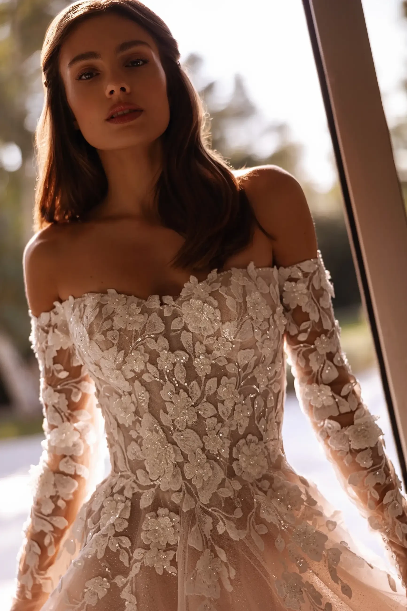 A woman wearing an elegant off-shoulder wedding dress with intricate floral embroidery and long lace sleeves, standing near a window with soft light illuminating her.