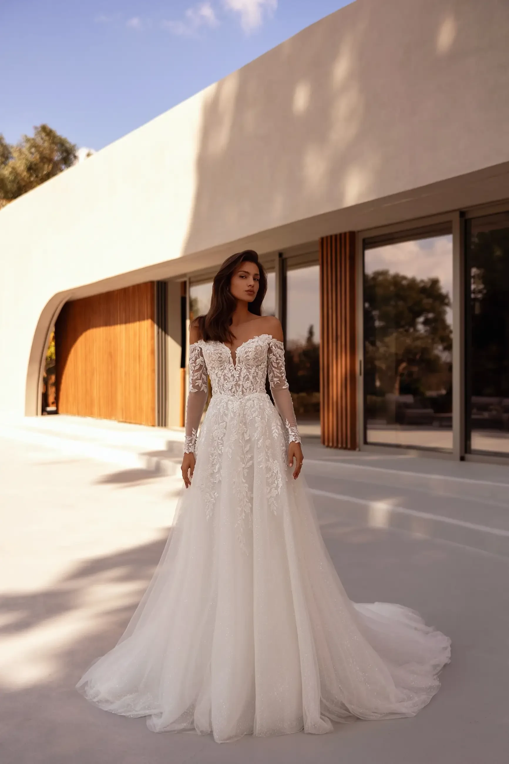 A woman stands outside a modern building, wearing an elegant off-shoulder lace wedding gown. The scene is bathed in soft sunlight, creating a serene ambiance.