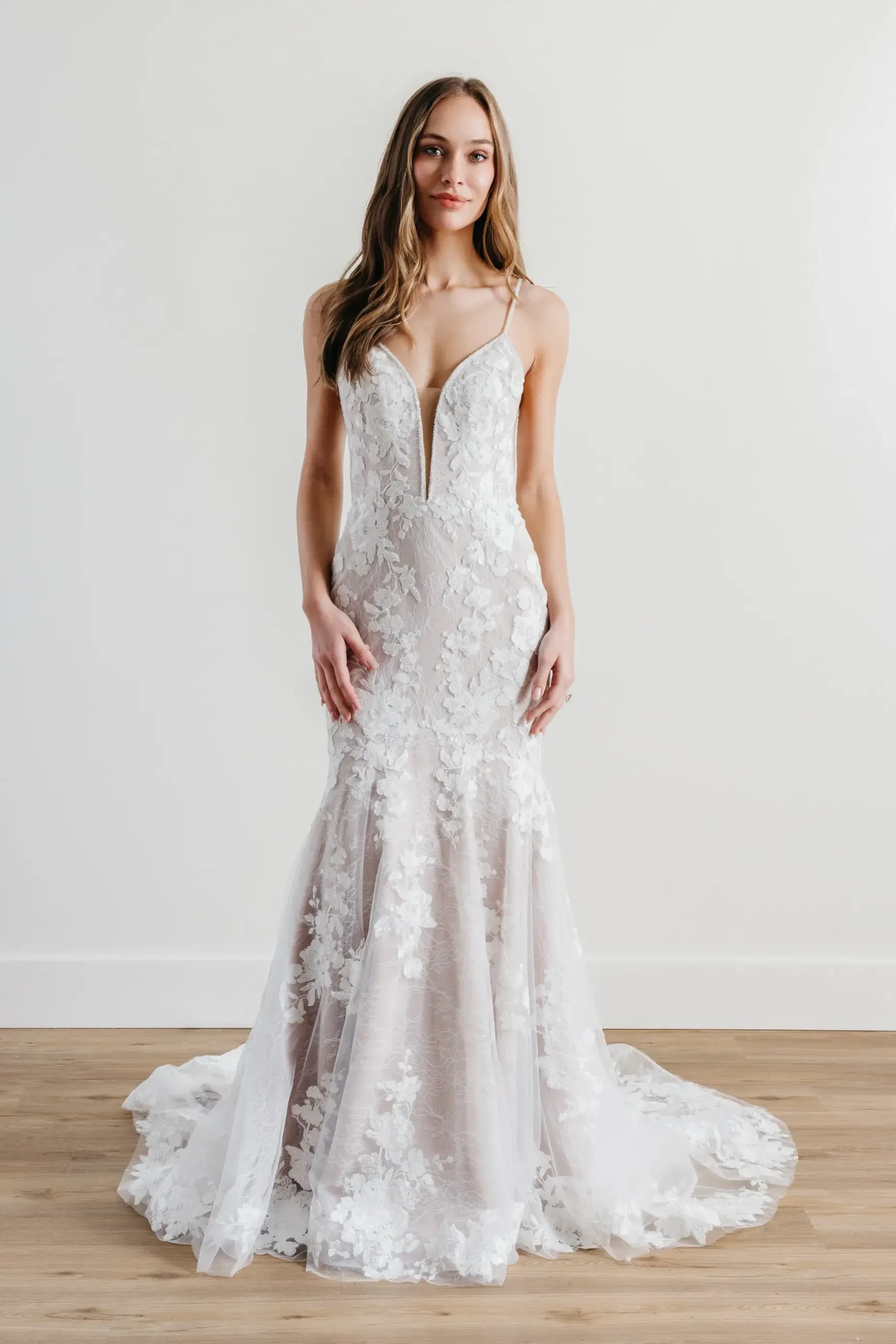 A woman stands in a fitted, sleeveless wedding gown with floral lace details and a deep V-neckline. The train flows elegantly on a wooden floor.