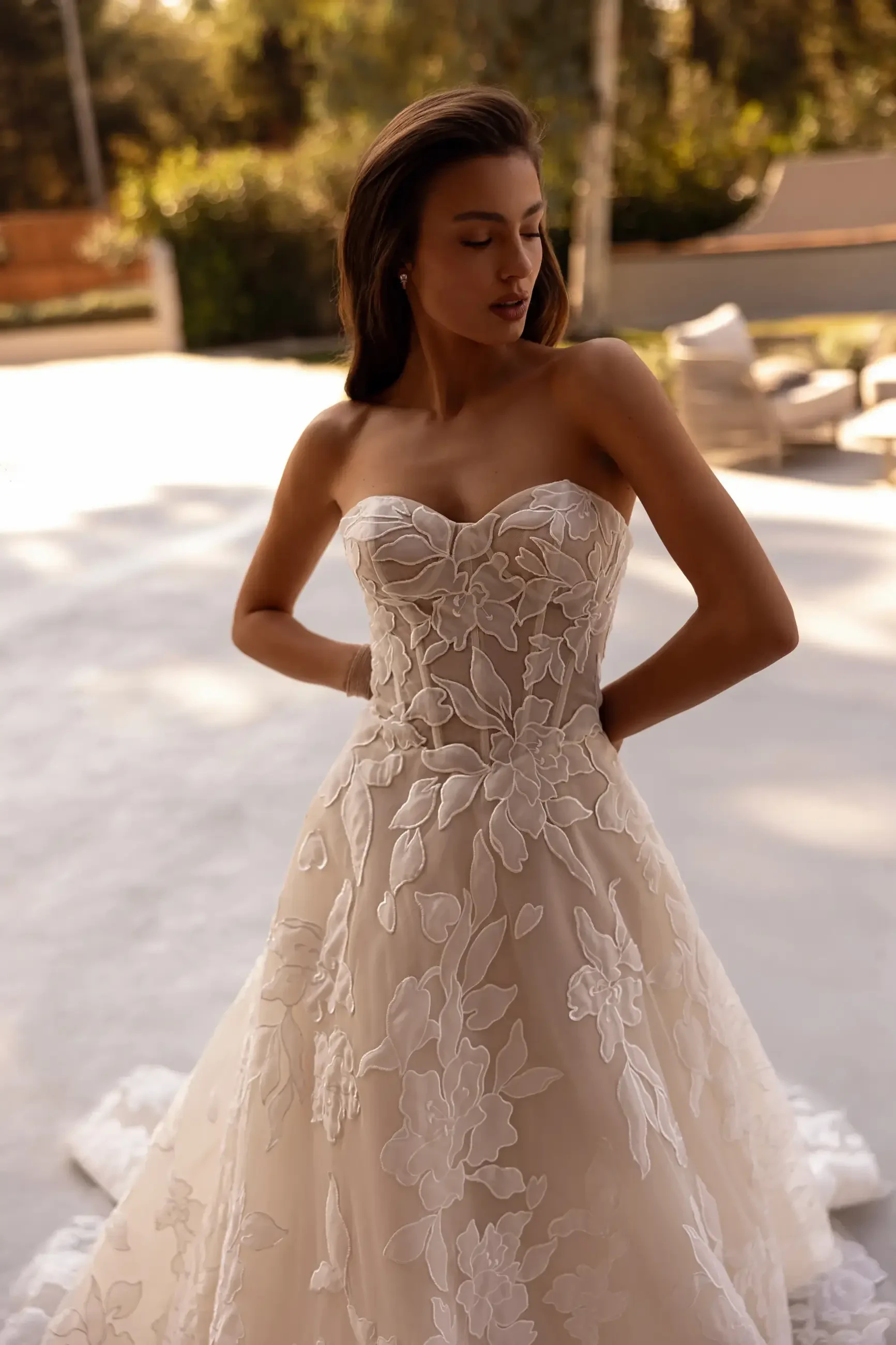 A woman wearing a strapless wedding dress with floral lace detailing, standing outdoors with a serene expression.