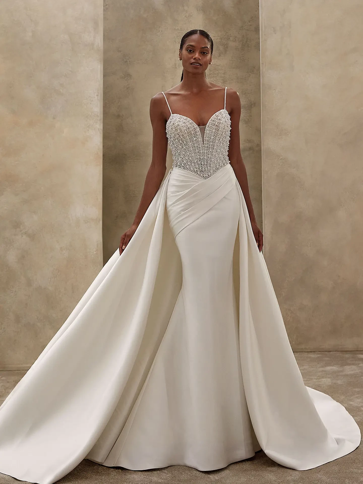 Elegant woman wearing a white wedding gown with a beaded bodice and a wide, flowing skirt. The background is a textured, light beige wall.