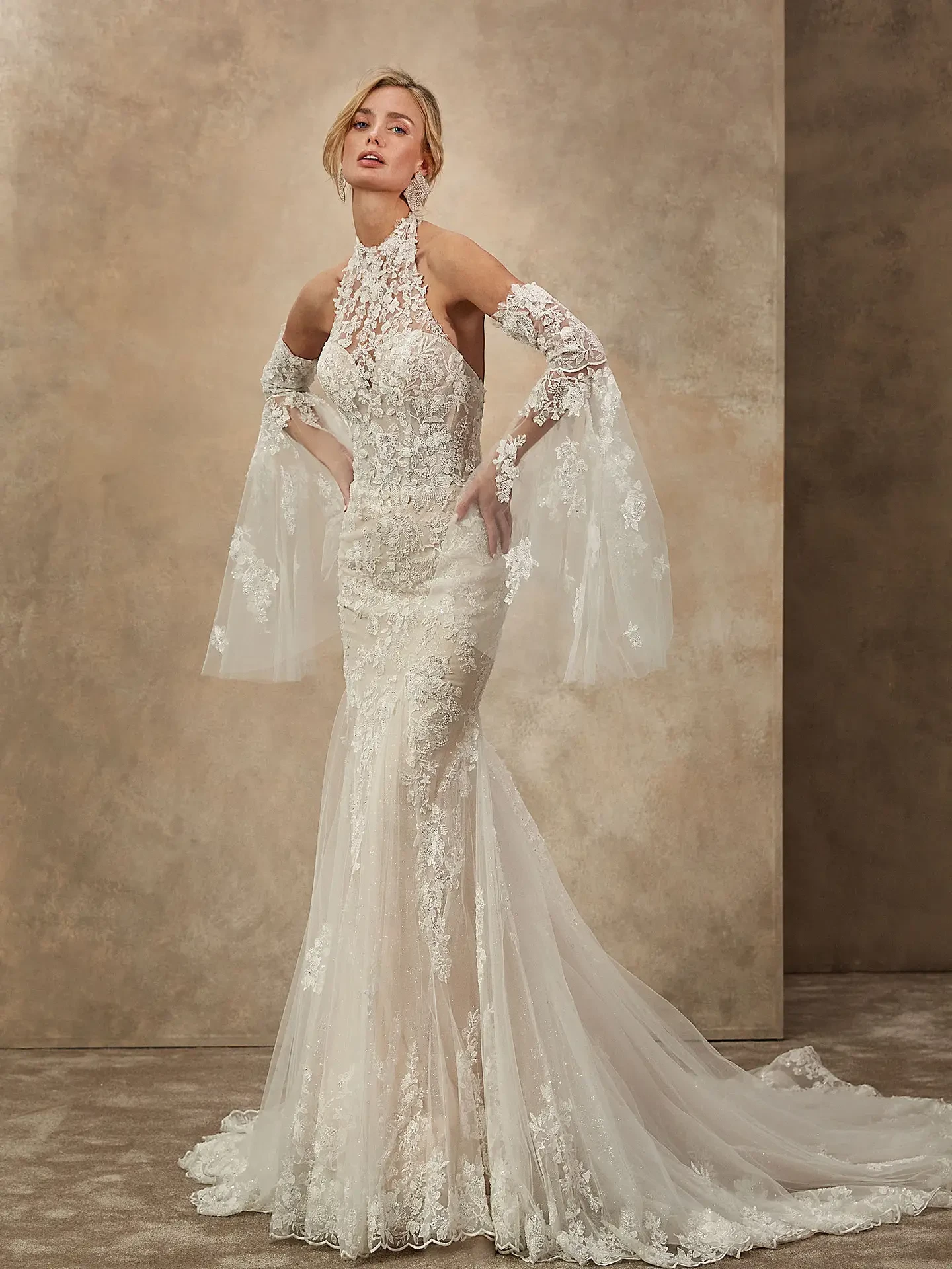 A woman poses in an elegant ivory lace wedding gown with a high neckline and sheer, flowing sleeves. The background is a neutral, textured wall.