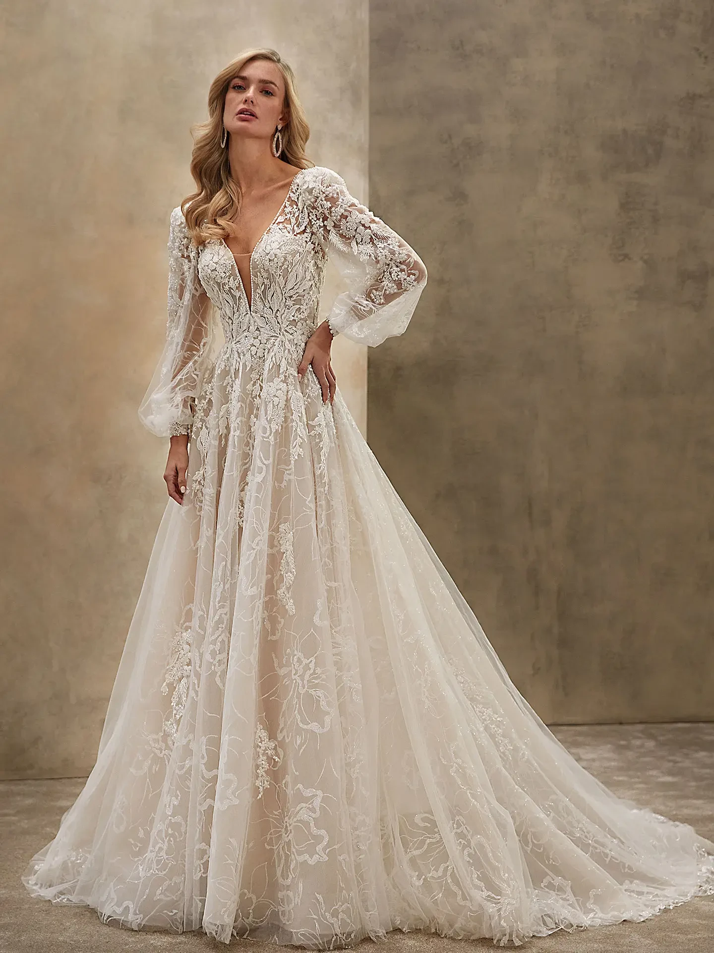 A woman in an elegant, lace wedding gown with sheer, billowy sleeves and floral patterns stands confidently against a textured, neutral backdrop.