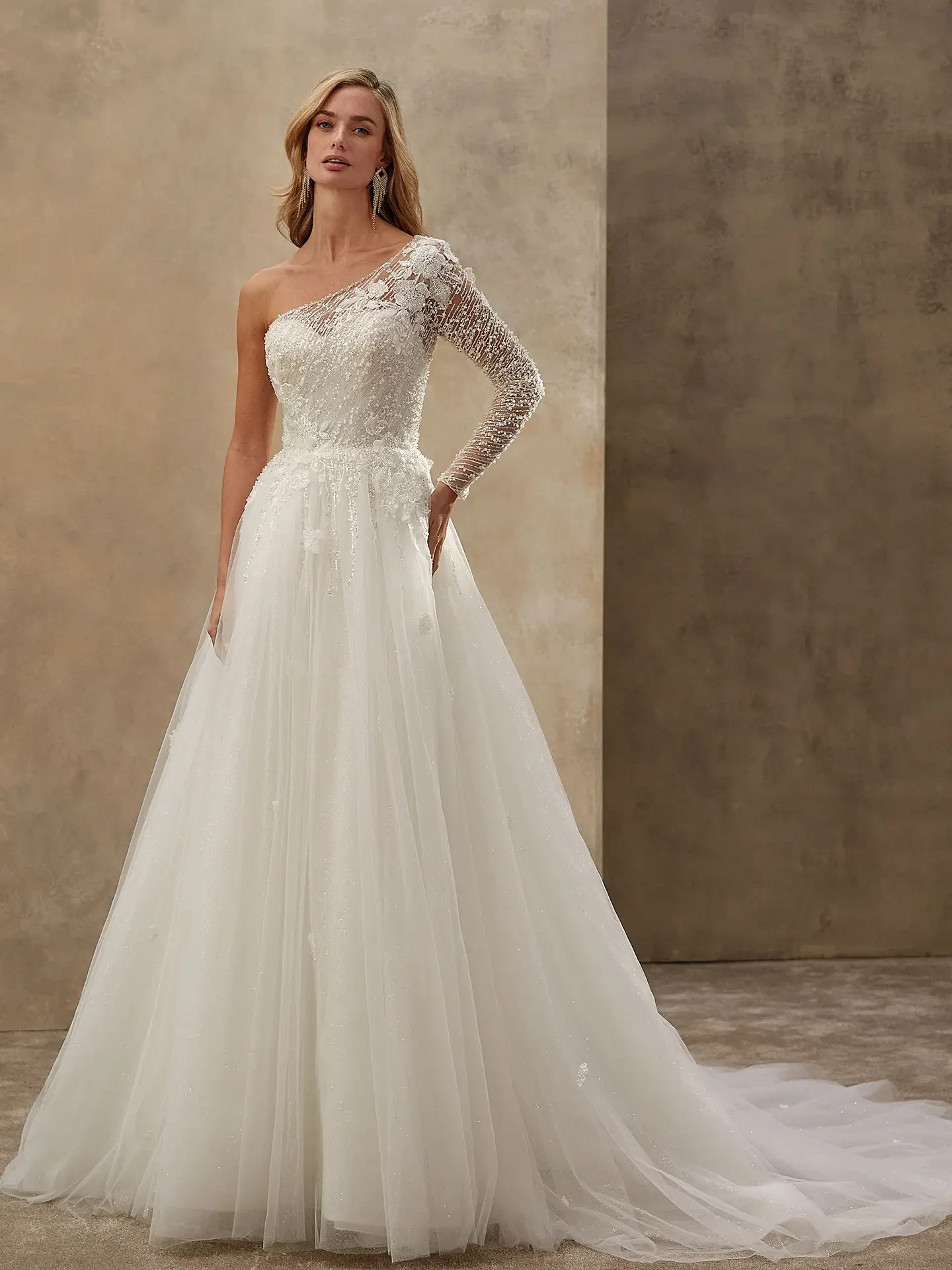 A woman wears an elegant, off-shoulder wedding dress with one lace sleeve. The dress features intricate floral detailing, set against a neutral background.