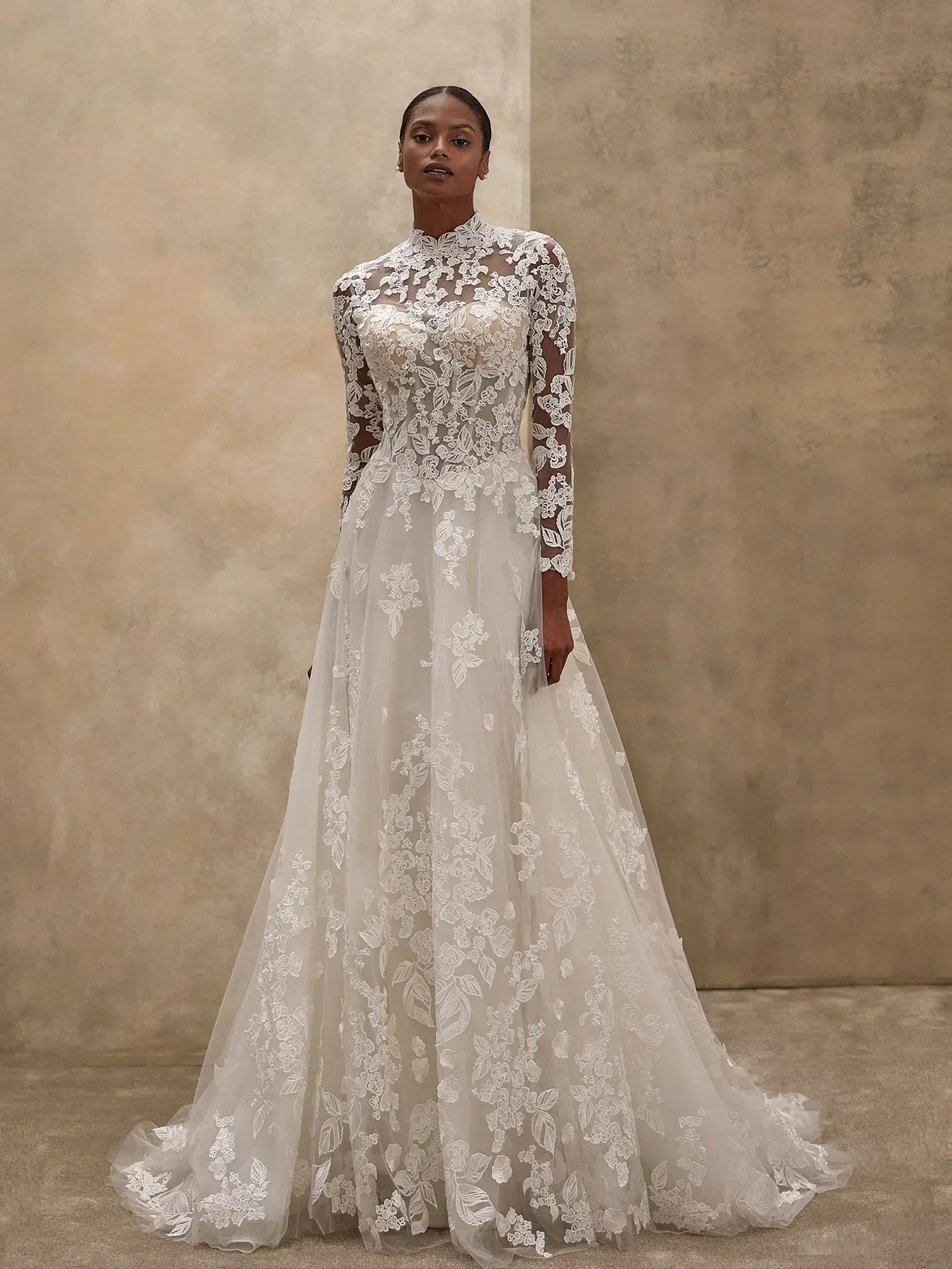 A woman stands gracefully in a modest, elegant white lace bridal gown with long sleeves and a high neck. The dress features intricate floral patterns. The background is a soft, beige wall, adding a serene and sophisticated tone.