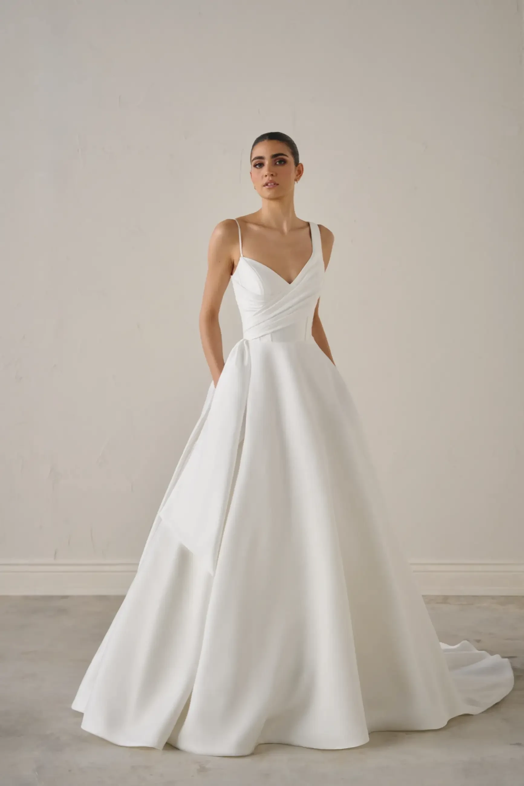 A model wearing a white gown with a fitted bodice and an A-line skirt, standing in a minimalist studio setting.