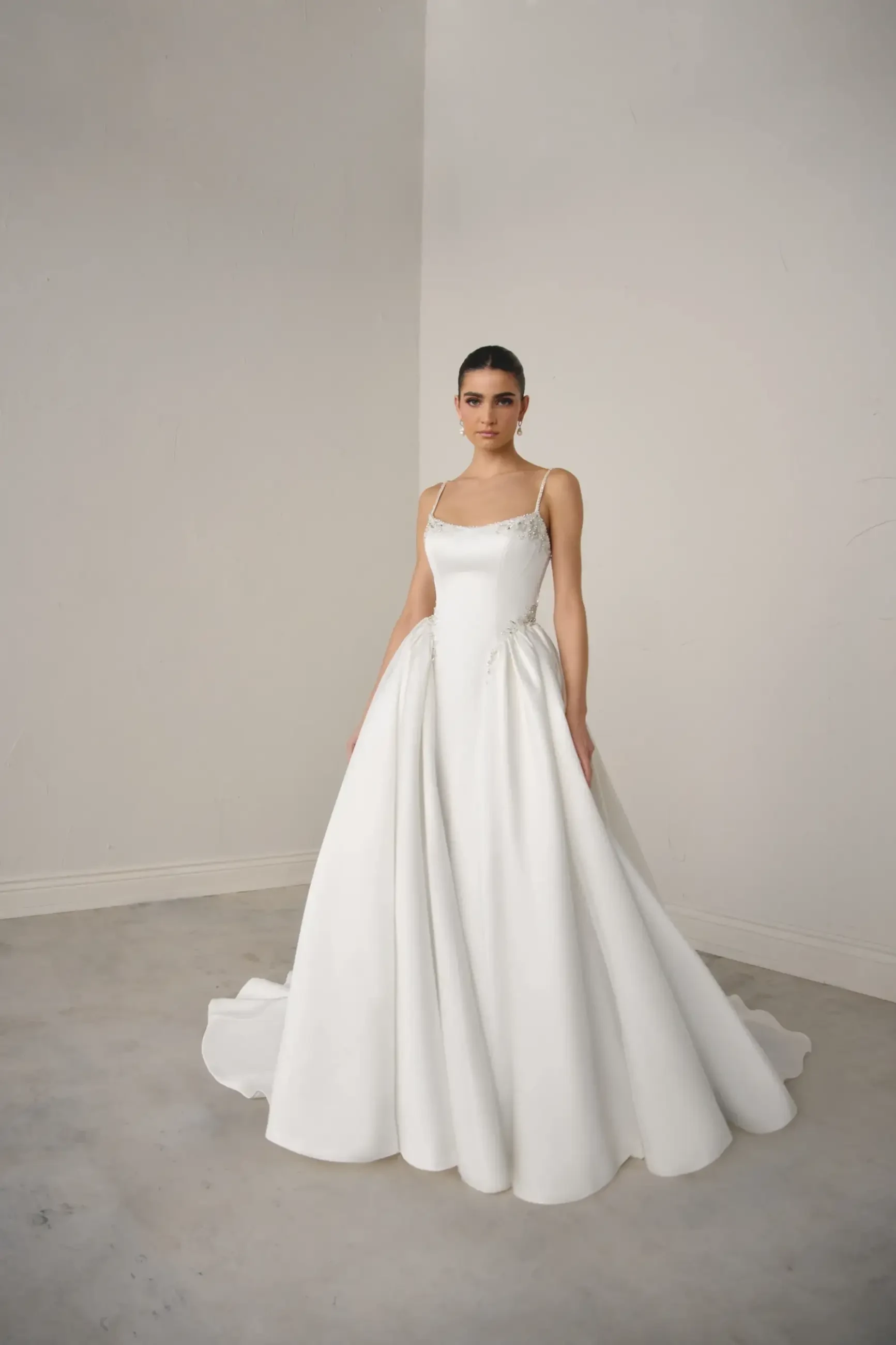 A model wearing a long, white gown with a fitted bodice and flowing skirt, standing in a minimalist setting with neutral colors.