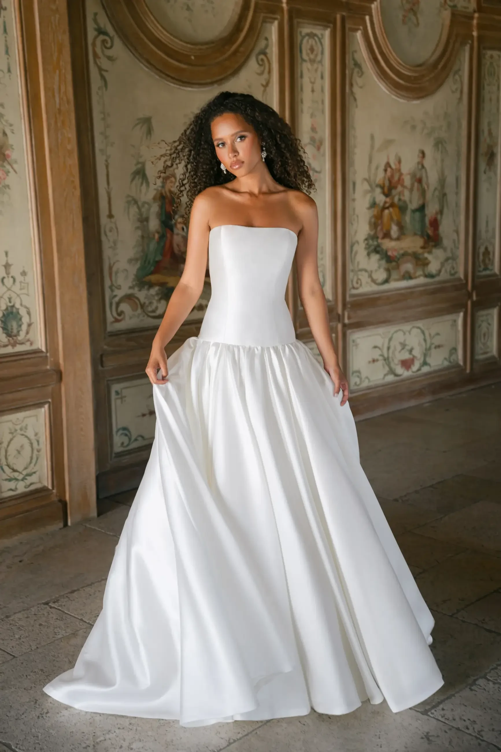 A woman wearing a strapless white gown stands gracefully in a beautifully decorated room with ornate walls.