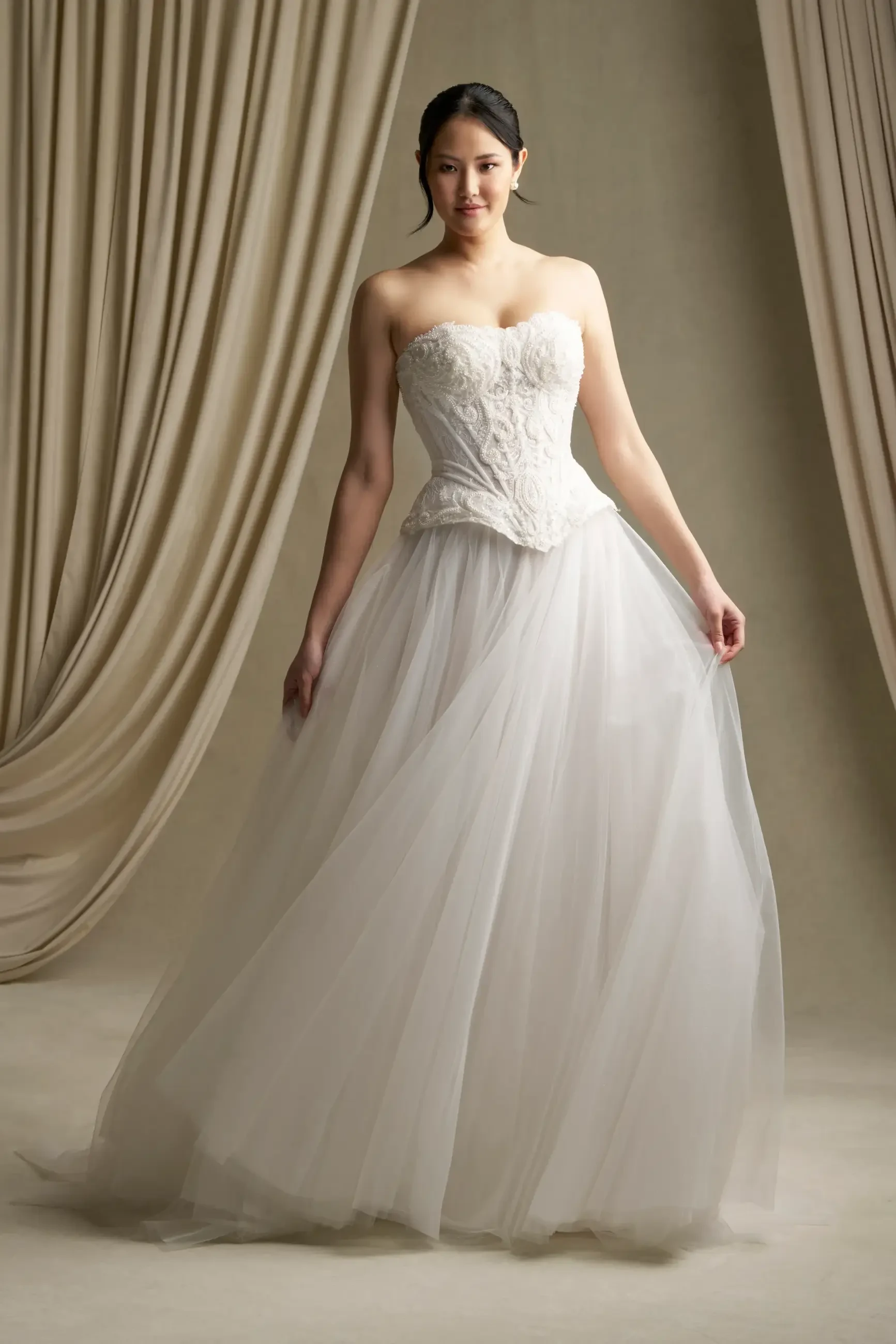 A woman wearing a strapless white wedding gown with a full tulle skirt, standing on display against a draped background.