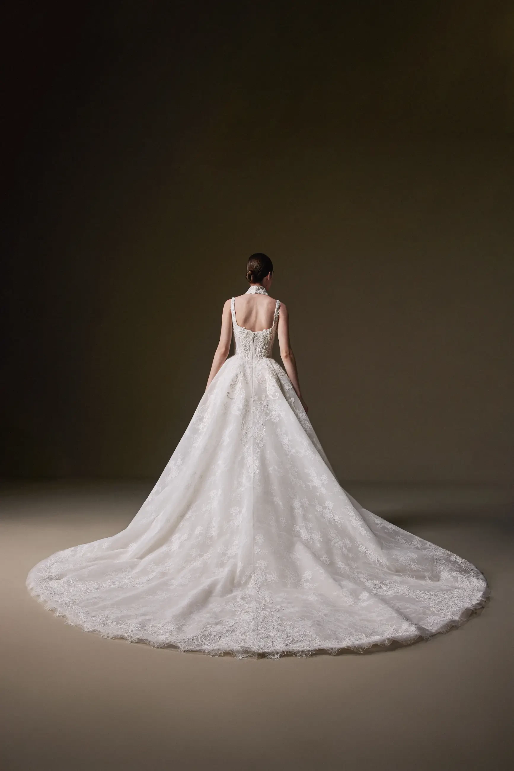 Back view of a bride in an elegant, white, lace wedding gown with a voluminous, flowing train, standing in a softly lit, neutral-toned room.