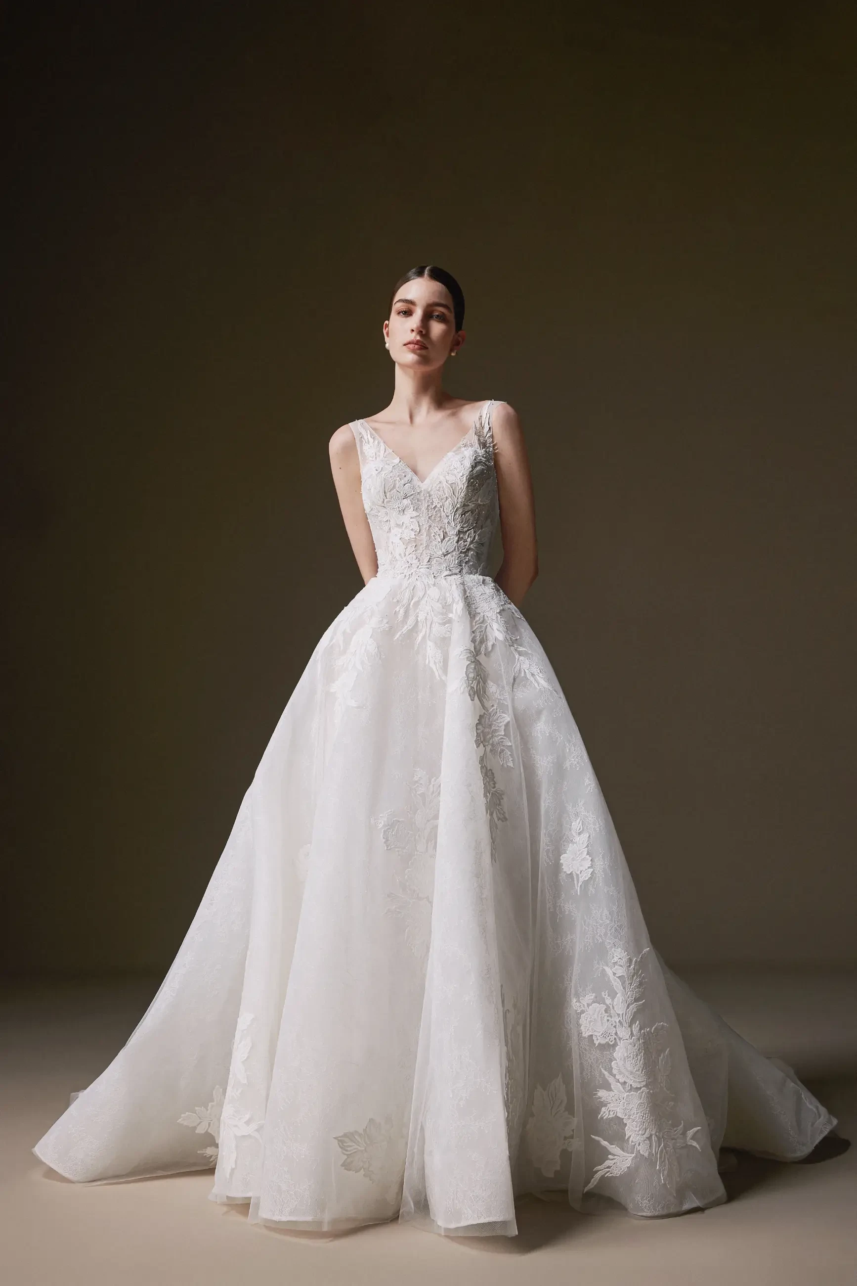 A woman in an elegant, voluminous white lace wedding dress stands in a neutral-toned studio. The setting is serene, emphasizing timeless beauty.
