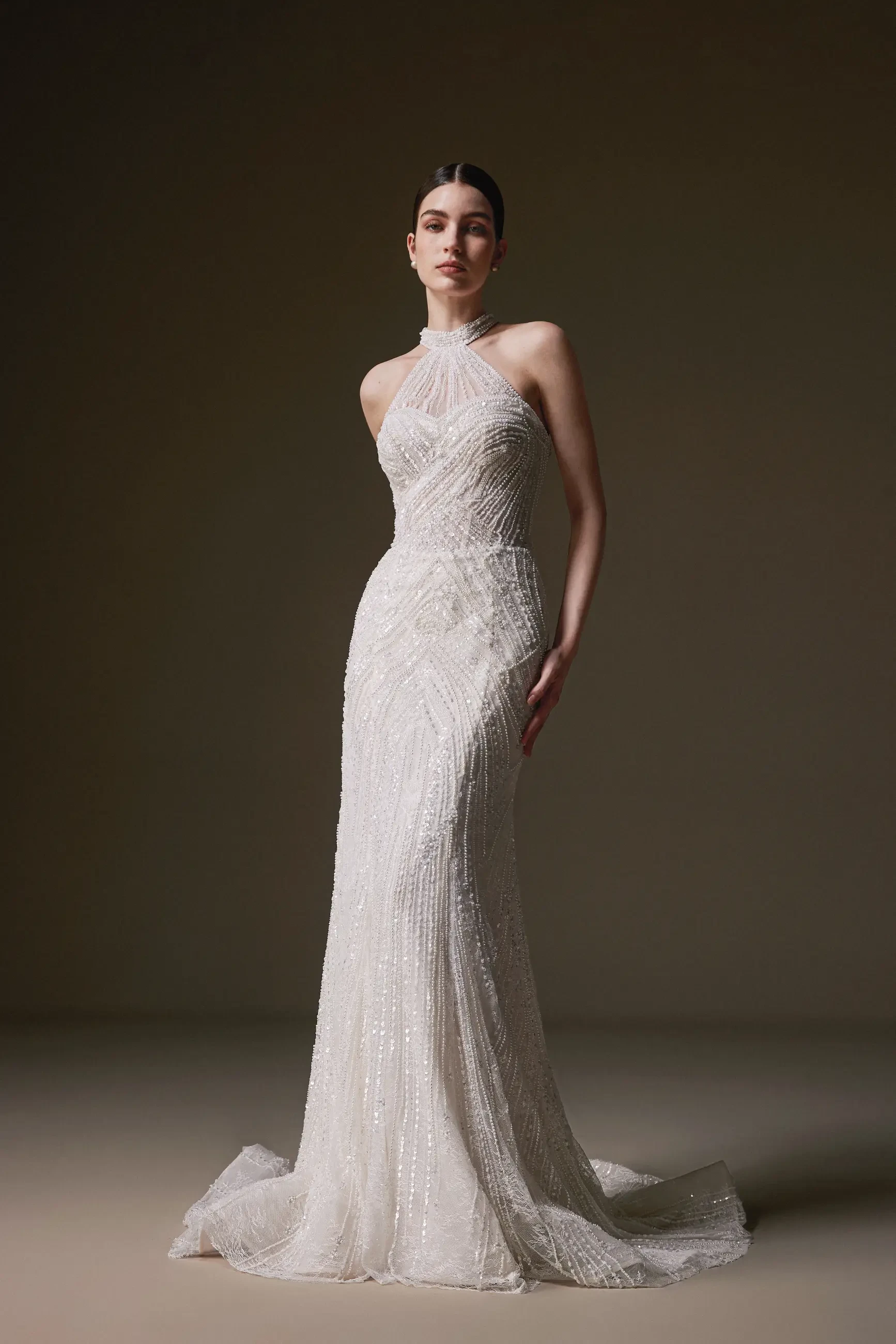 Elegant model wearing a shimmering, form-fitting, floor-length gown with a halter neckline, conveying sophistication and grace against a neutral backdrop.