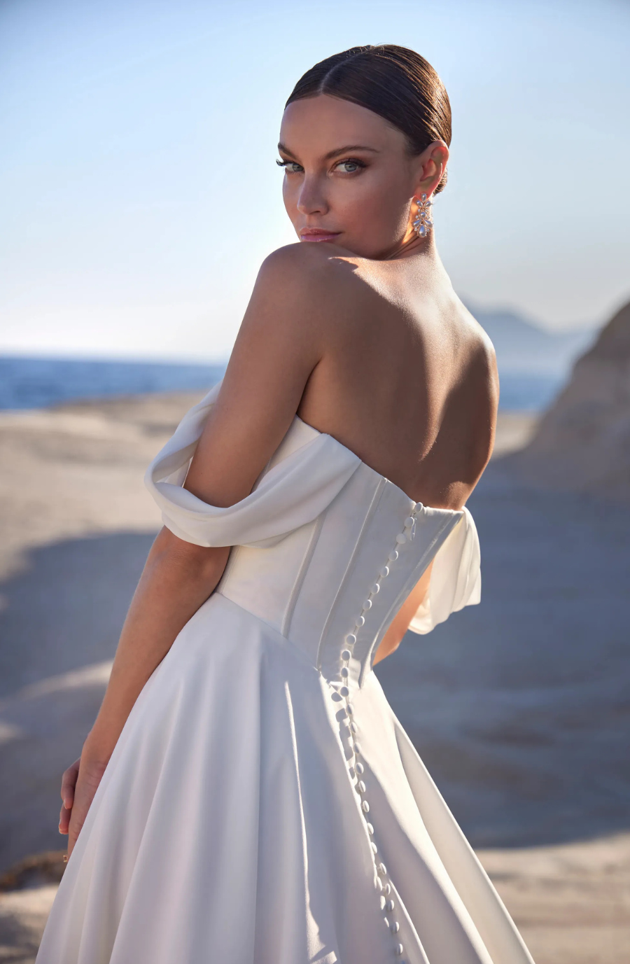 A woman in an off-shoulder wedding dress stands on a beach, gazing over her shoulder. The dress has button detailing, and the scene is serene and sunlit.