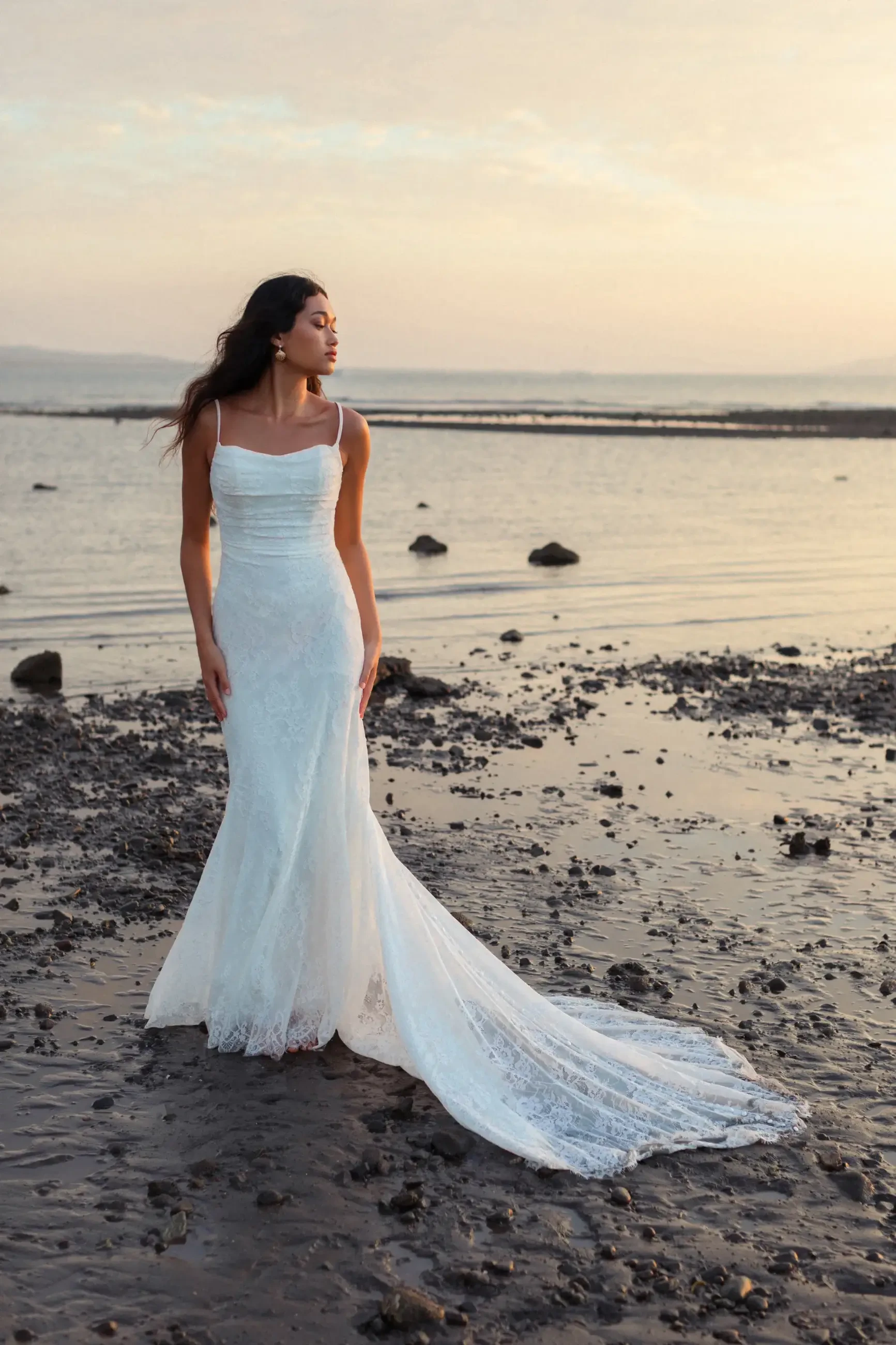 A woman in a flowing white gown stands on a rocky shore at sunset. She gazes to the side, exuding calm and elegance against the serene water backdrop.