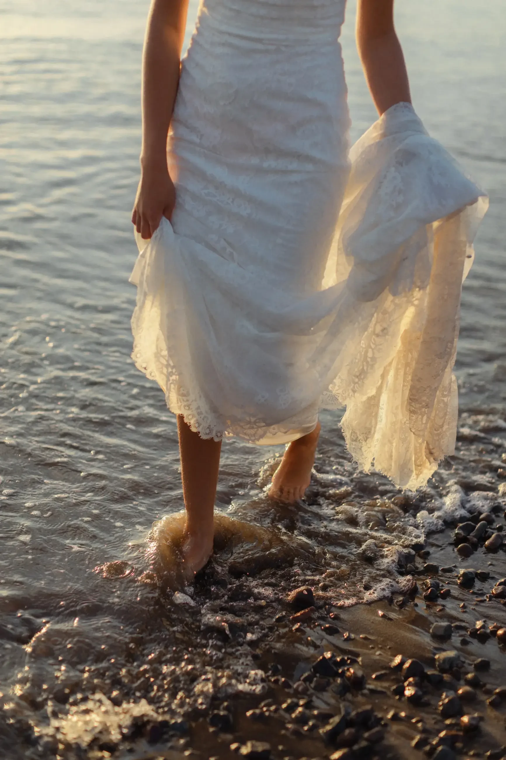 Bride in a white lace dress walks barefoot along a pebbled beach, with gentle waves lapping around her feet, evoking a serene, romantic atmosphere.