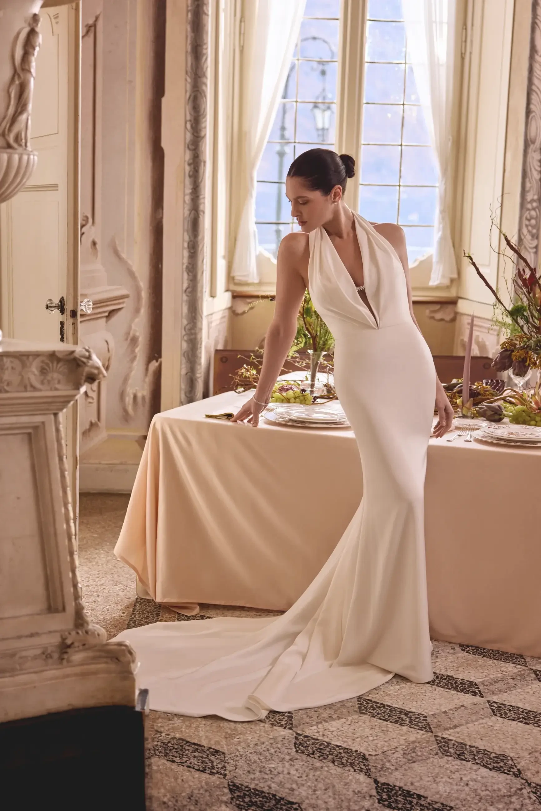 Elegant woman in a sleek white gown stands by a table with a beige cloth, in a vintage room with large windows, creating a serene and graceful atmosphere.