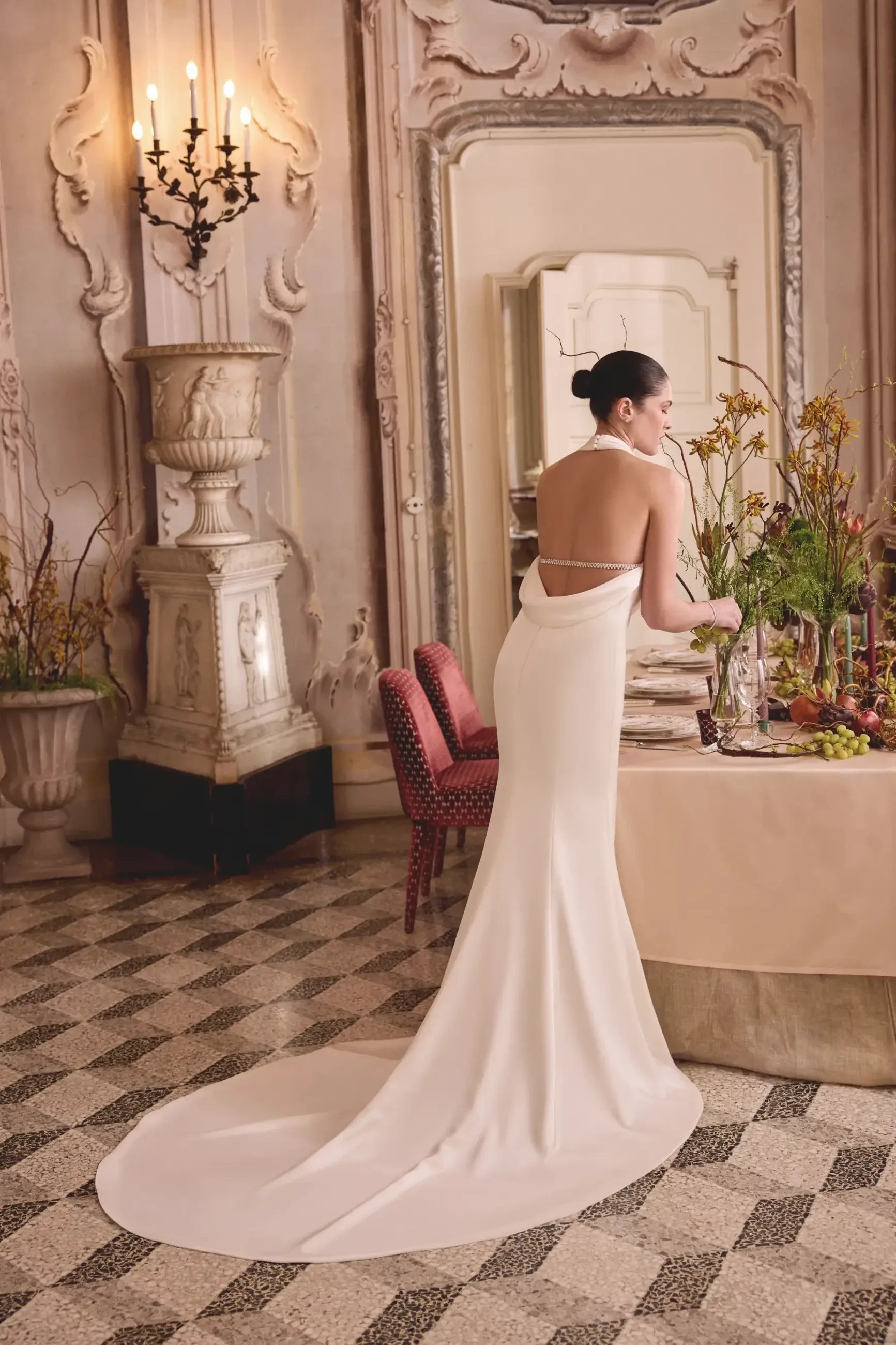 A woman in a backless white dress stands near a table adorned with flowers and food in an elegant indoor setting.
