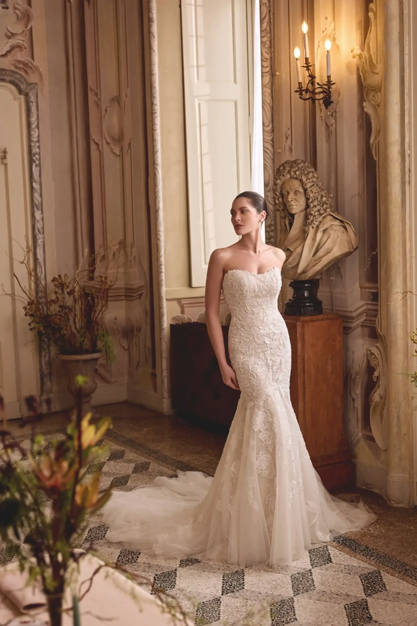 A woman wearing a strapless bridal gown stands elegantly in a beautifully decorated room with historical architecture and a bust statue nearby.