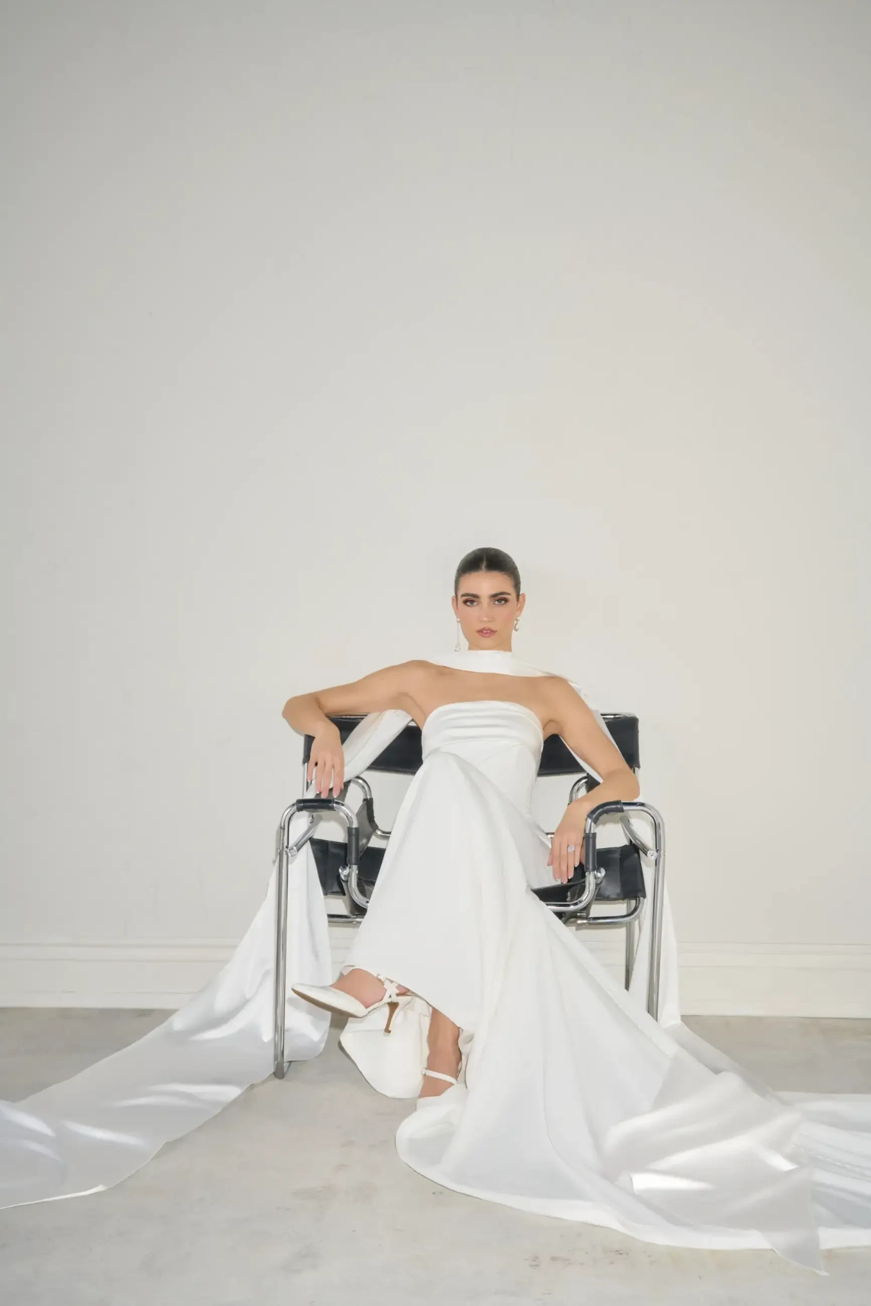 A woman in a flowing white gown sits confidently on a modern metal chair, against a plain background, conveying elegance and empowerment.