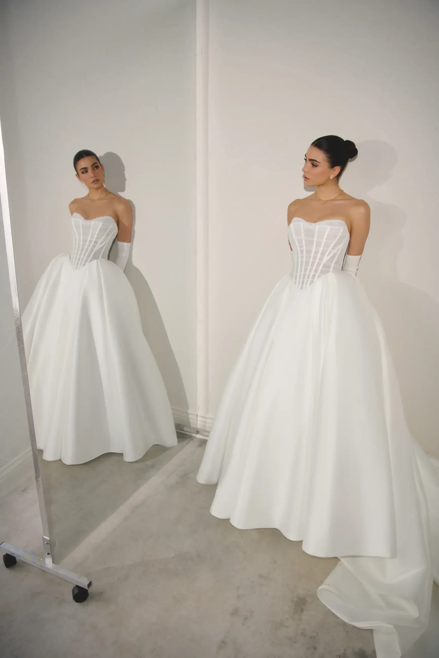 Bride in a strapless, white ball gown admires her reflection in a mirror. The elegant dress features a structured bodice and flowing skirt, evoking a serene, romantic mood.