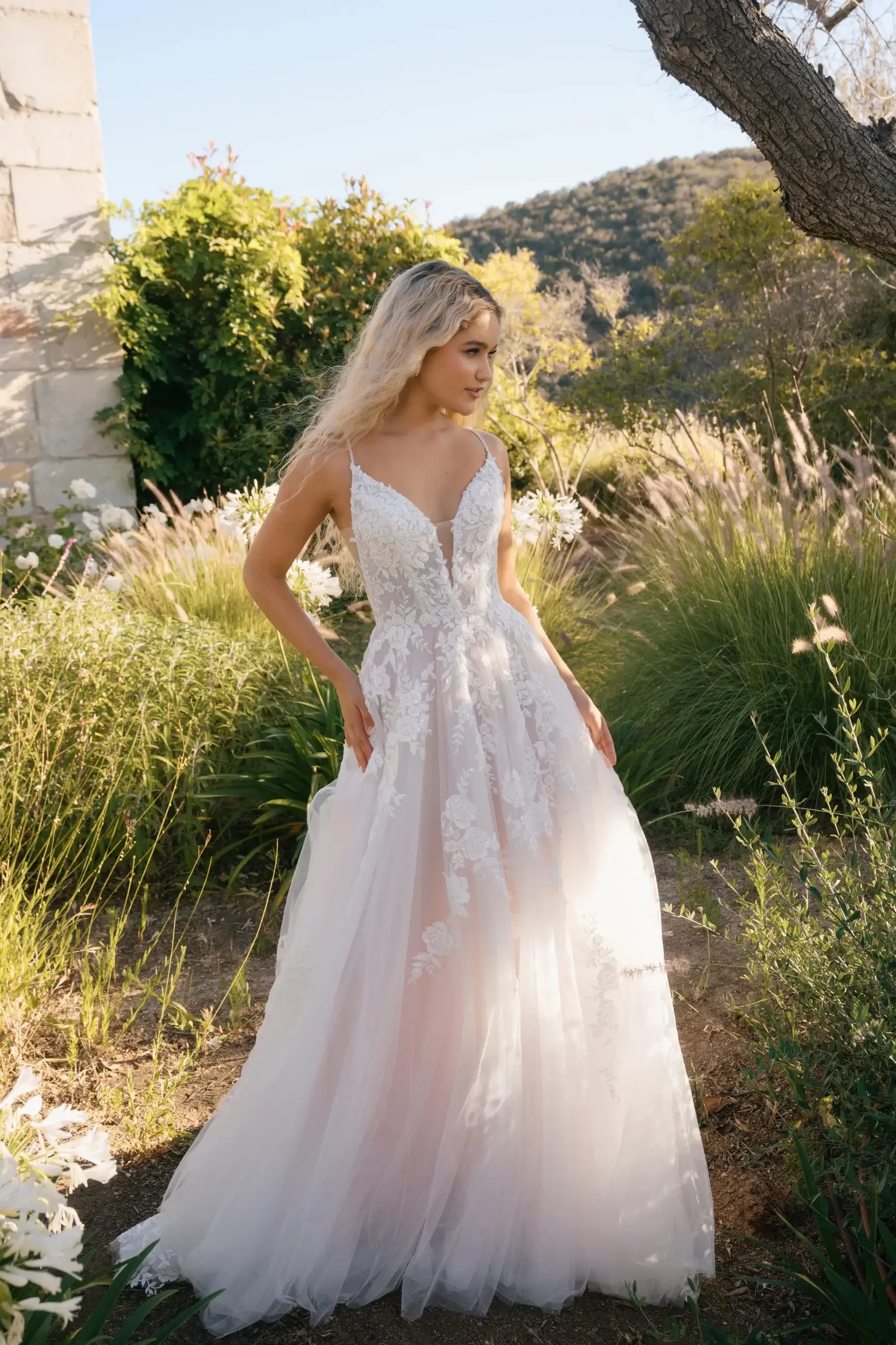 A woman in a flowing white lace wedding dress stands in a sunlit garden. The scenery includes greenery and distant hills, creating a serene, romantic mood.
