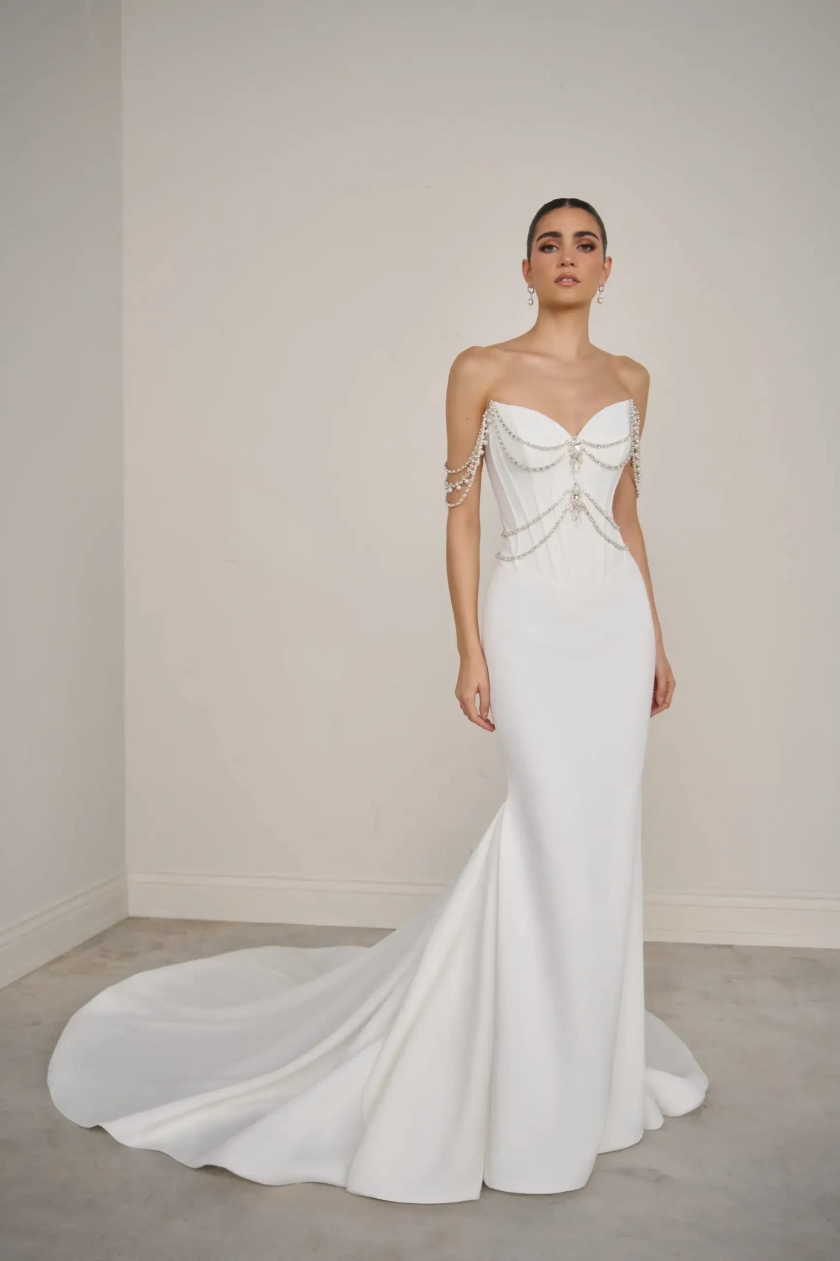 A model wearing a sleek, white evening gown with off-the-shoulder straps and decorative chain details. The dress has a fitted silhouette and a flowing train.