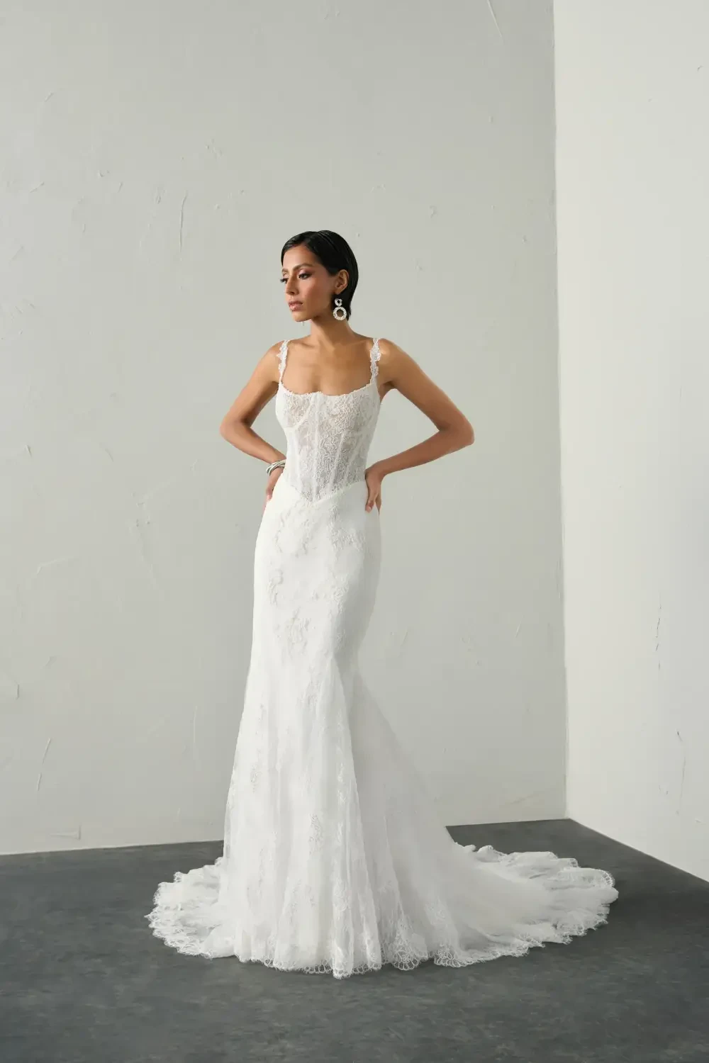 A woman in a fitted white lace wedding dress with a train, posed against a plain background.