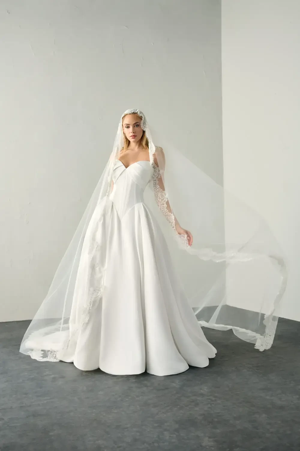 A model wearing a white wedding dress with a long veil, standing against a neutral backdrop.