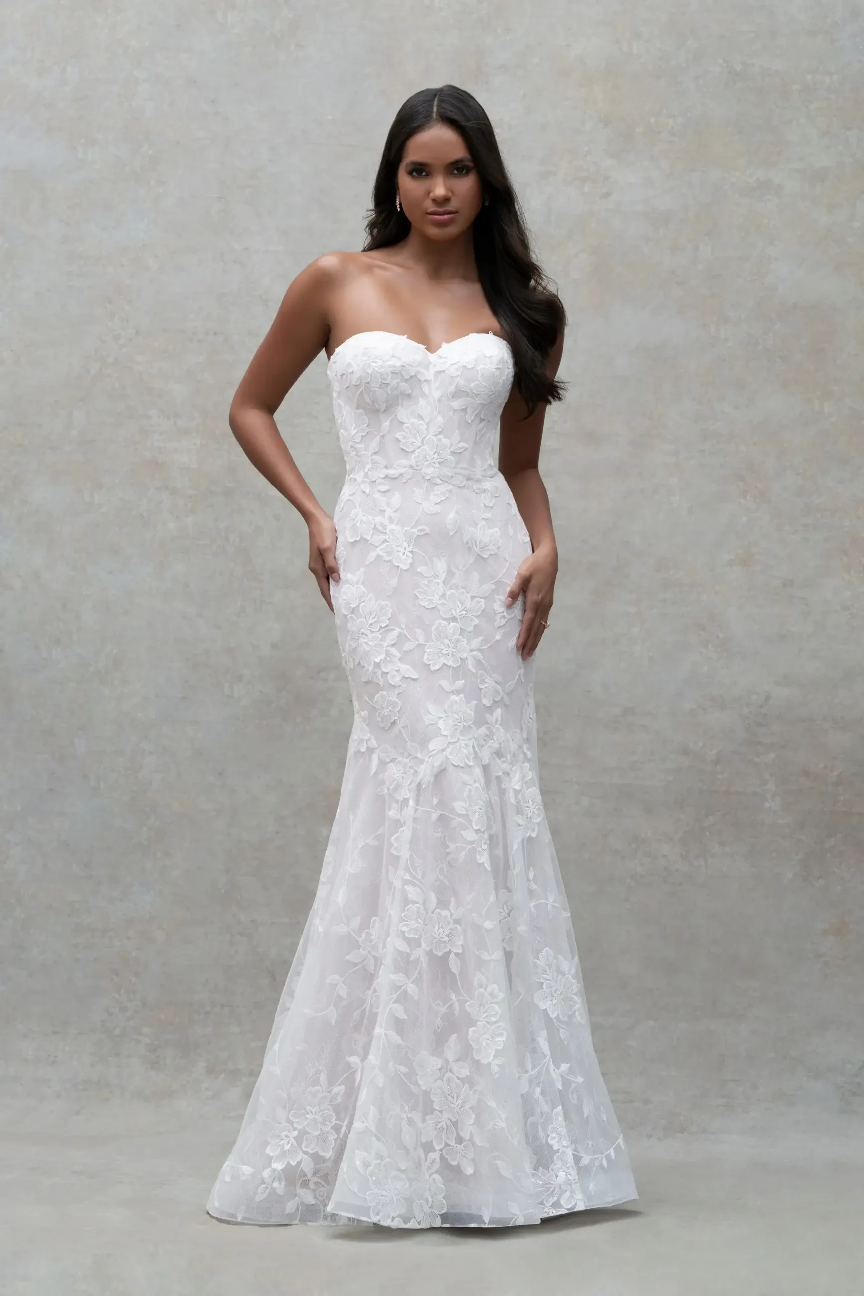 A woman is wearing a strapless white dress with a fitted silhouette and lace detailing, posing in front of a neutral background.